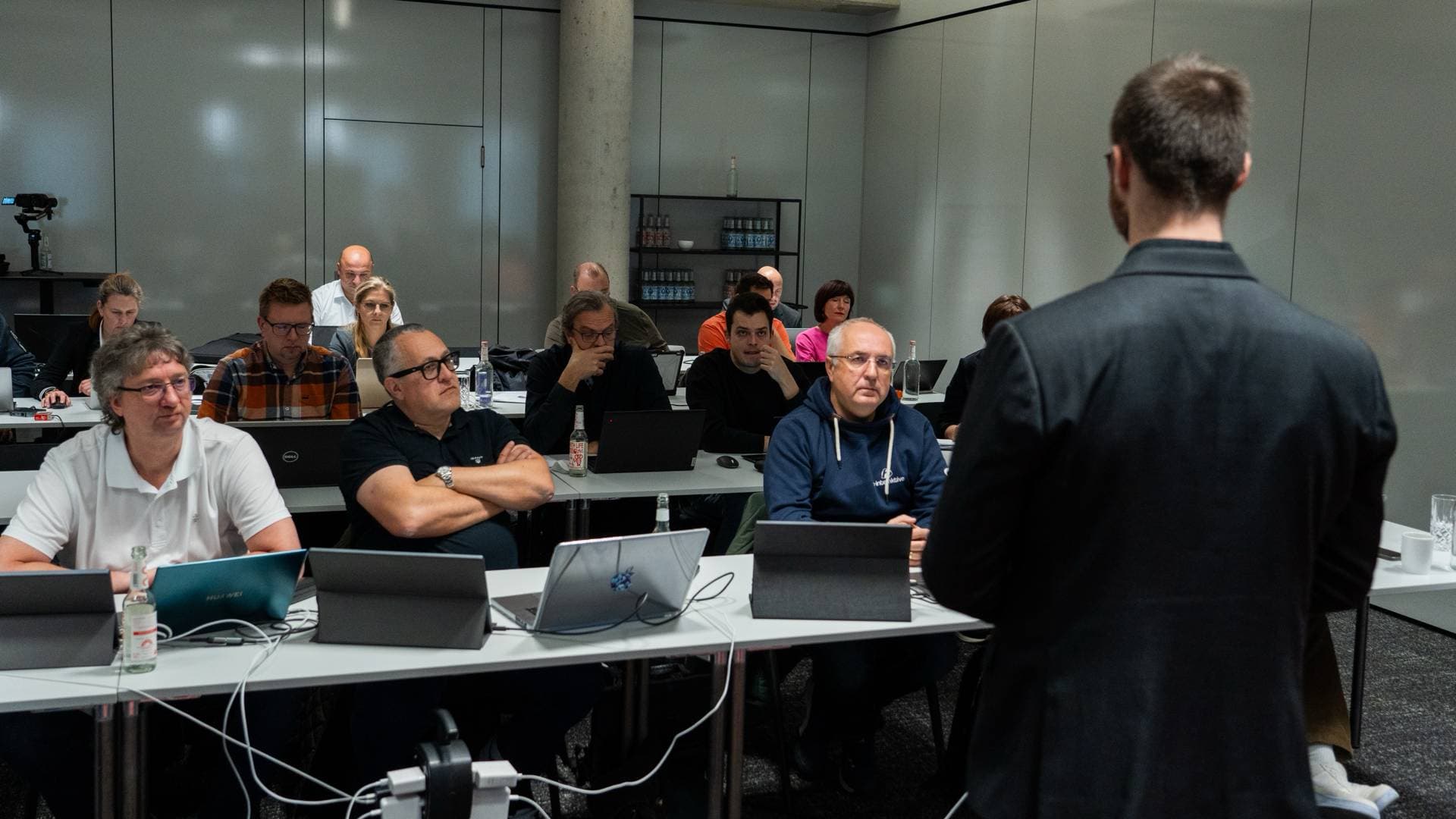 Teilnehmer eines KI-Workshops sitzen mit Laptops an Tischen und hören dem Trainer zu
